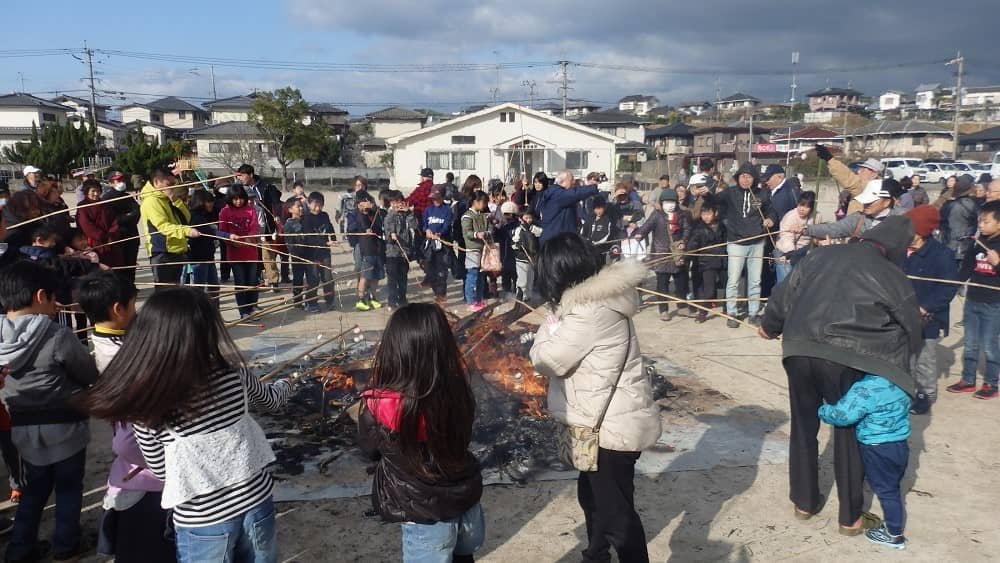 残り火で子どもたちや地域の方が竹さおの先に餅をつるし焼いている写真